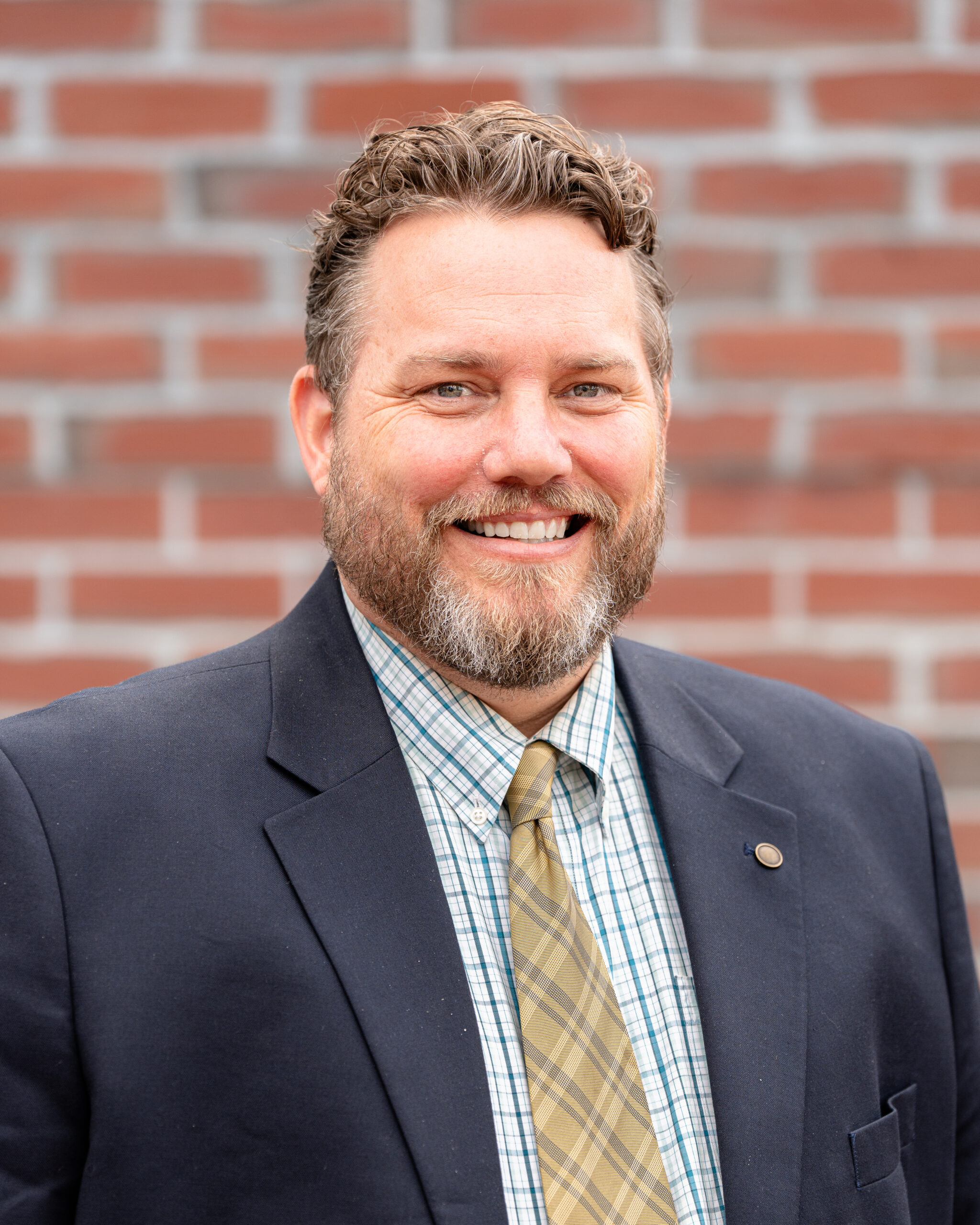 Atlee is a smiling middle aged man with short brown hair and a short beard, both starting to turn gray. He is wearing a blue suit jacket, blue and white shirt and a yellow tie.