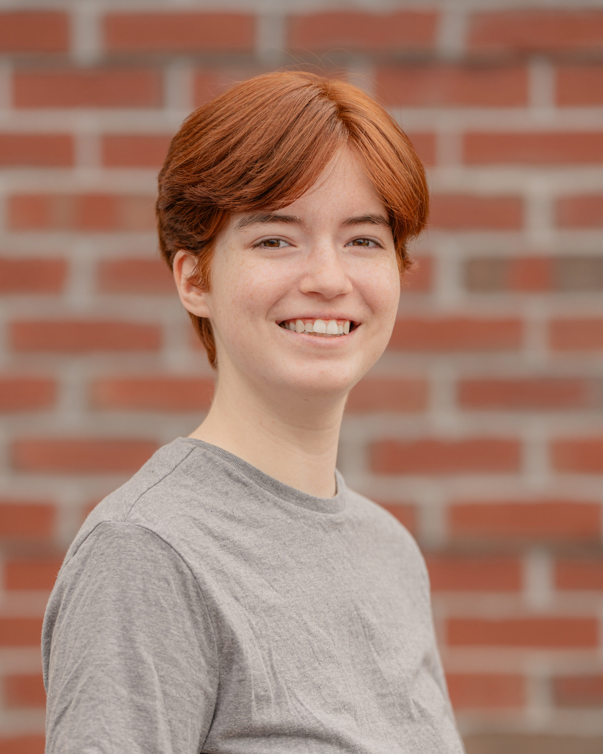 Aydan has short red hair and wears a brown t-shirt. They stand smiling in front of a brick wall.