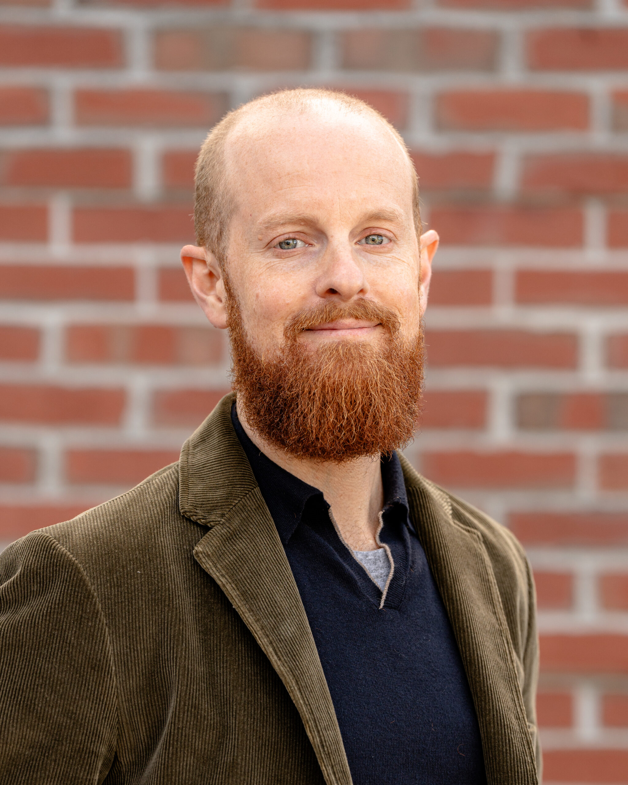 Barrett, a white male with a red beard and blue eyes. He wears a green blazer and black sweater.