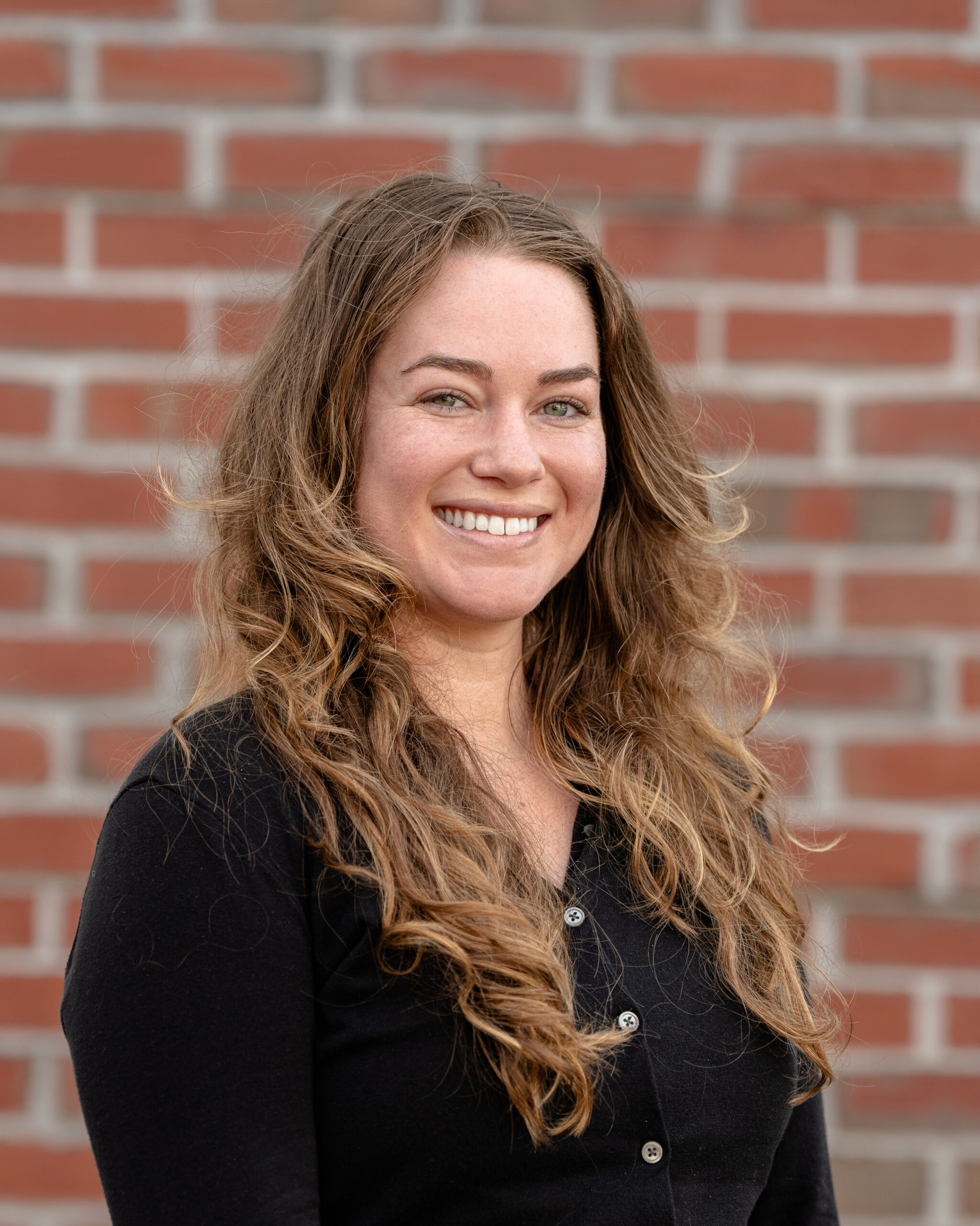 Bridget, a white woman with long brown hair smiles in front of a brick wall.