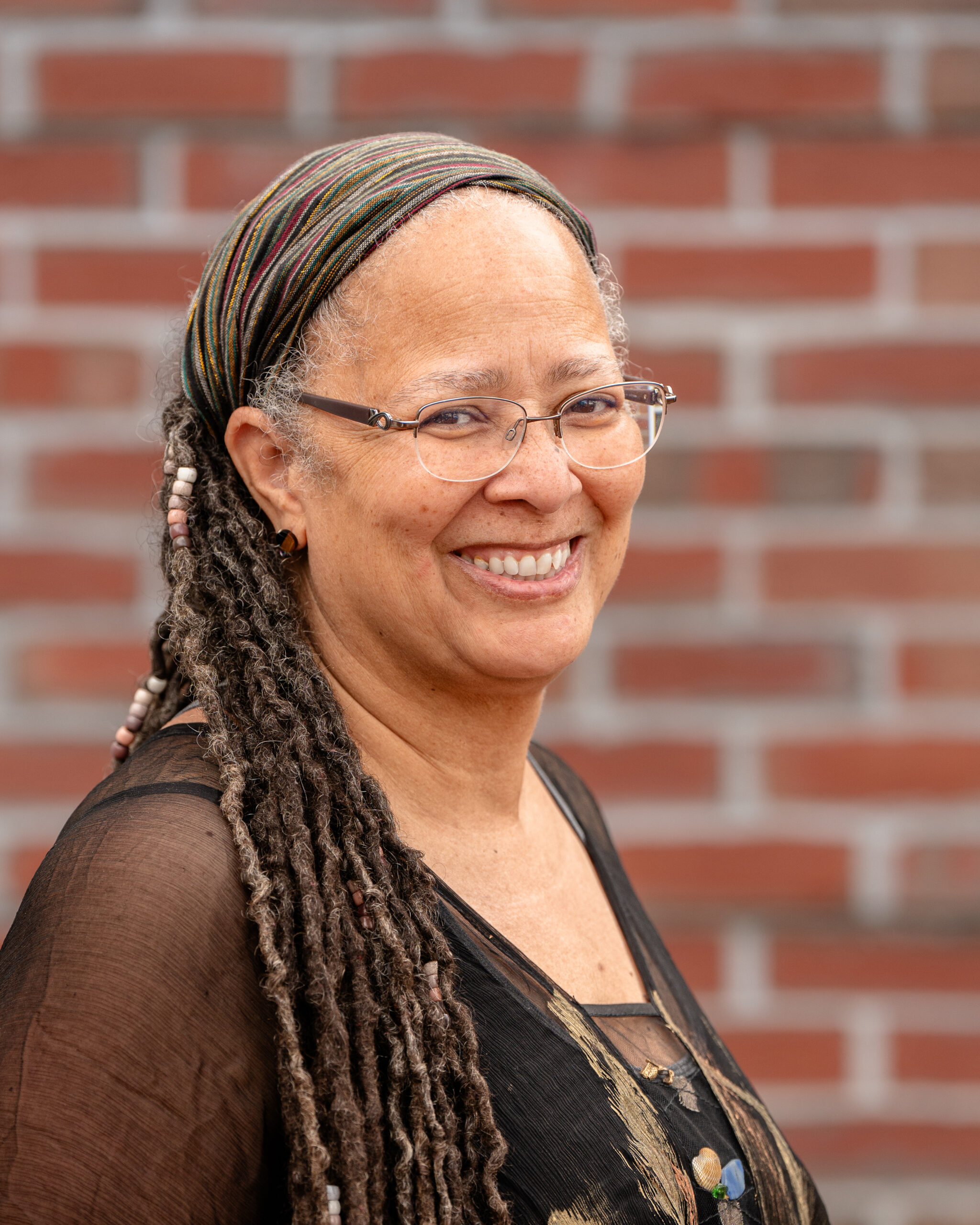 Denise has long black braids and wears a headband, black top and glasses.