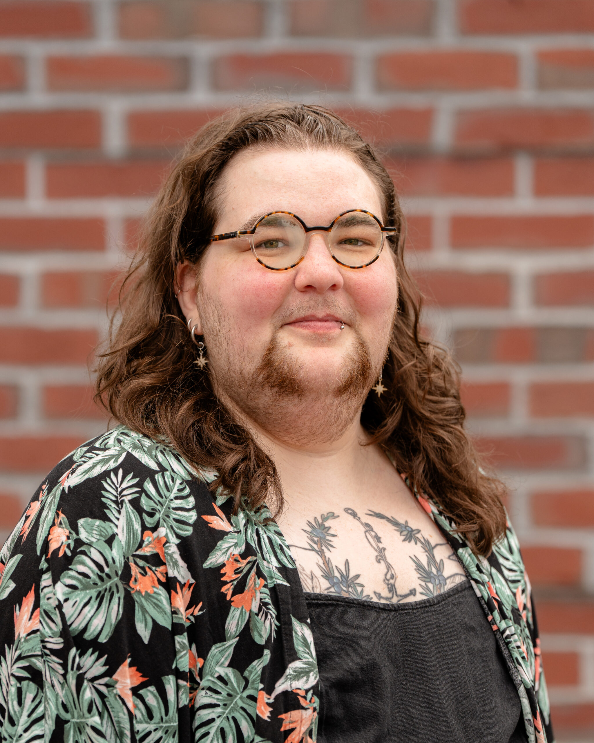 Harper, a white person with long wavy brown hair, a beard, and brown glasses. They wear a floral shirt.