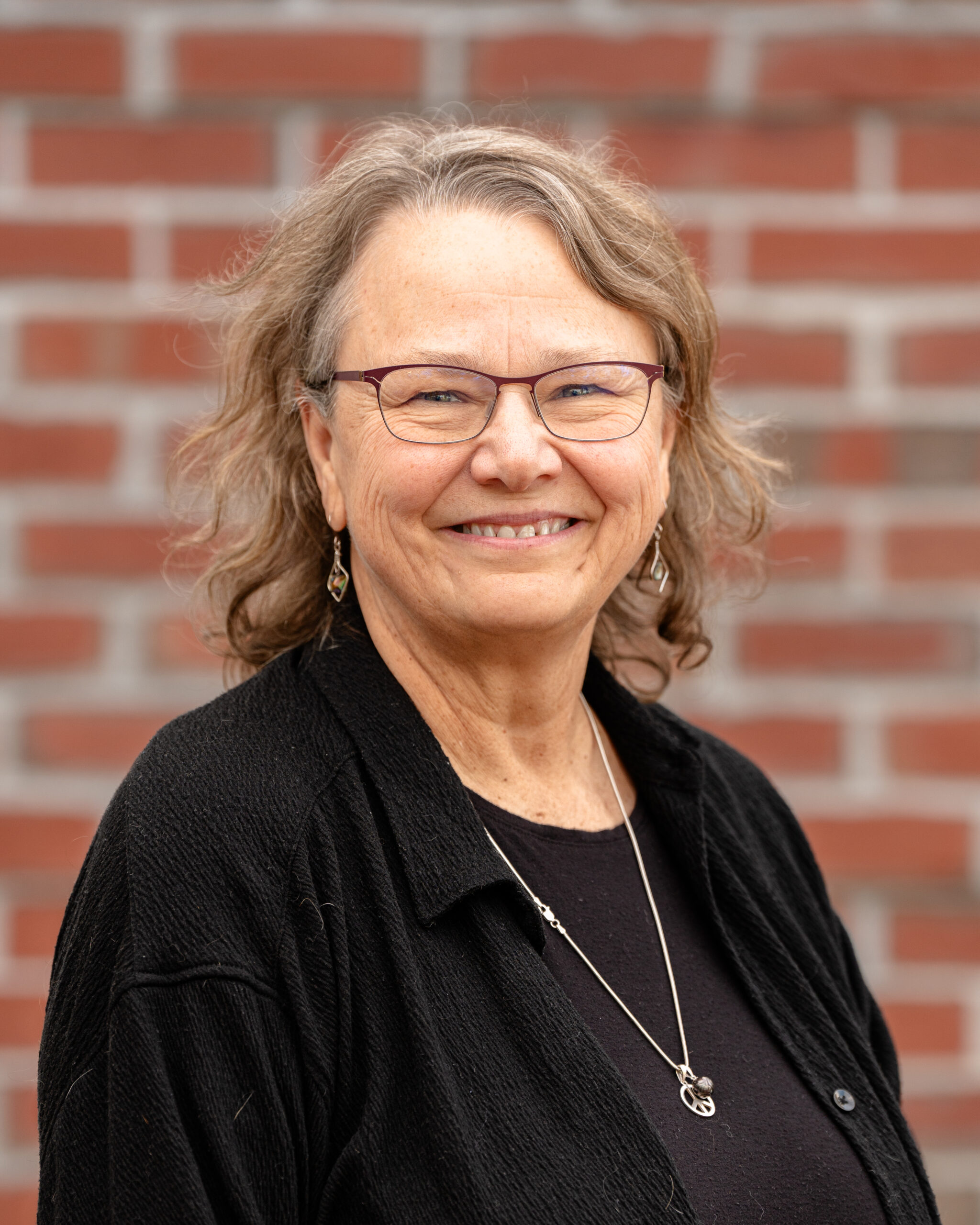 Jane is awhite woman, with glasses, brown medium length hair and a black shirt.