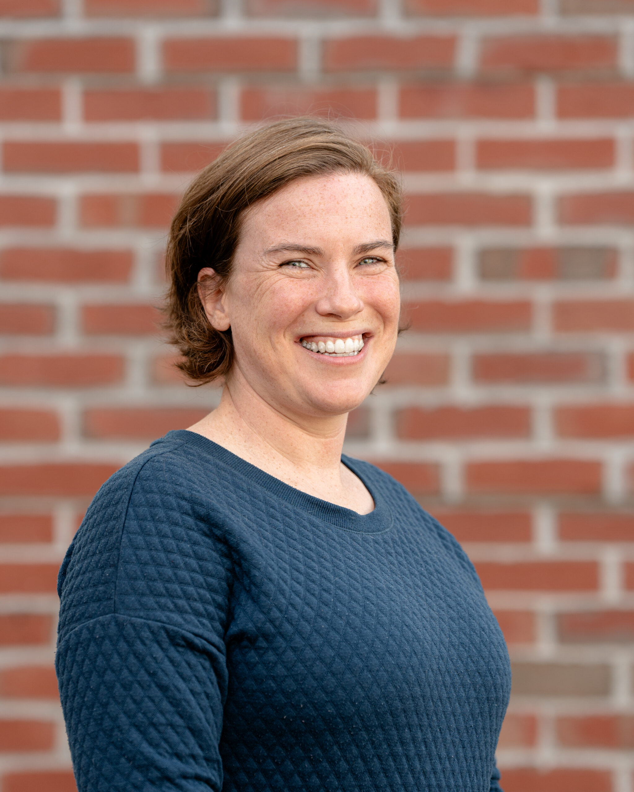 Jennifer, a white woman with brown hair and wearing a blue shirt, is standing in front of a brick wall. She is smiling at the camera.