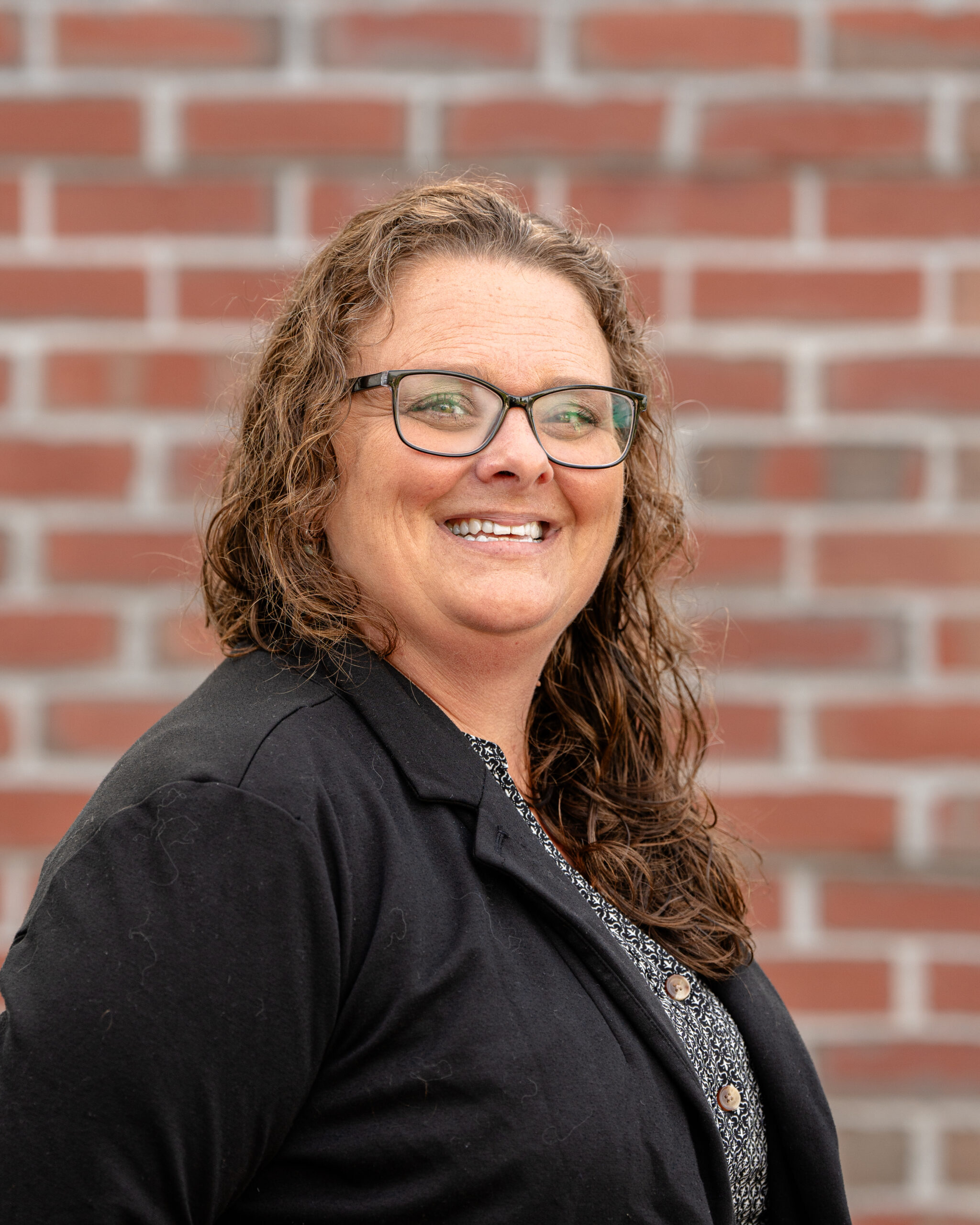 Katrina, a white woman with curly brown hair and brown glasses. She wears a black blazer and stands in front of a brick wall.