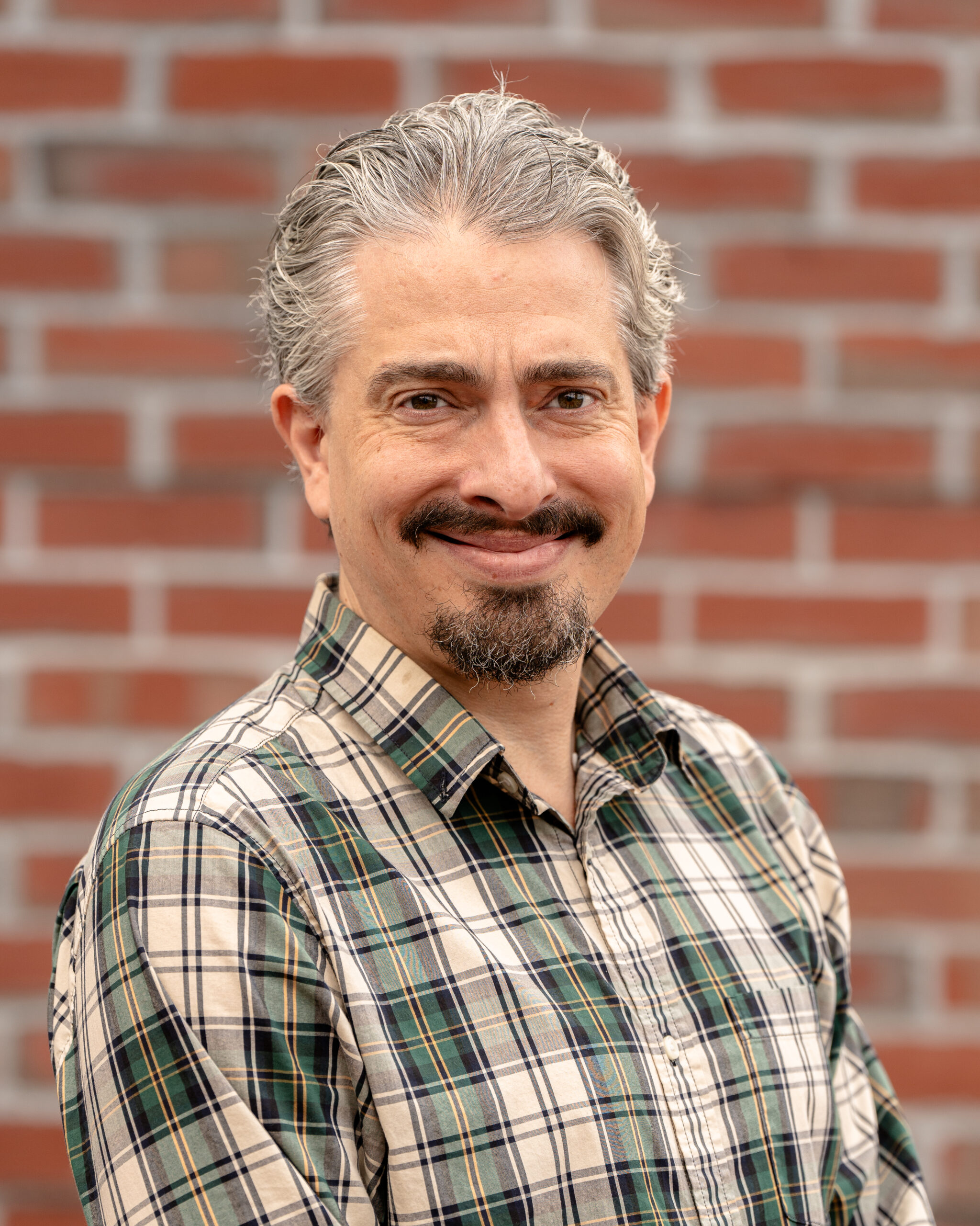 Kevin, a white man with short gray hair and a dark beard. He wears a green and white plaid shirt and stands in front of a brick wall.