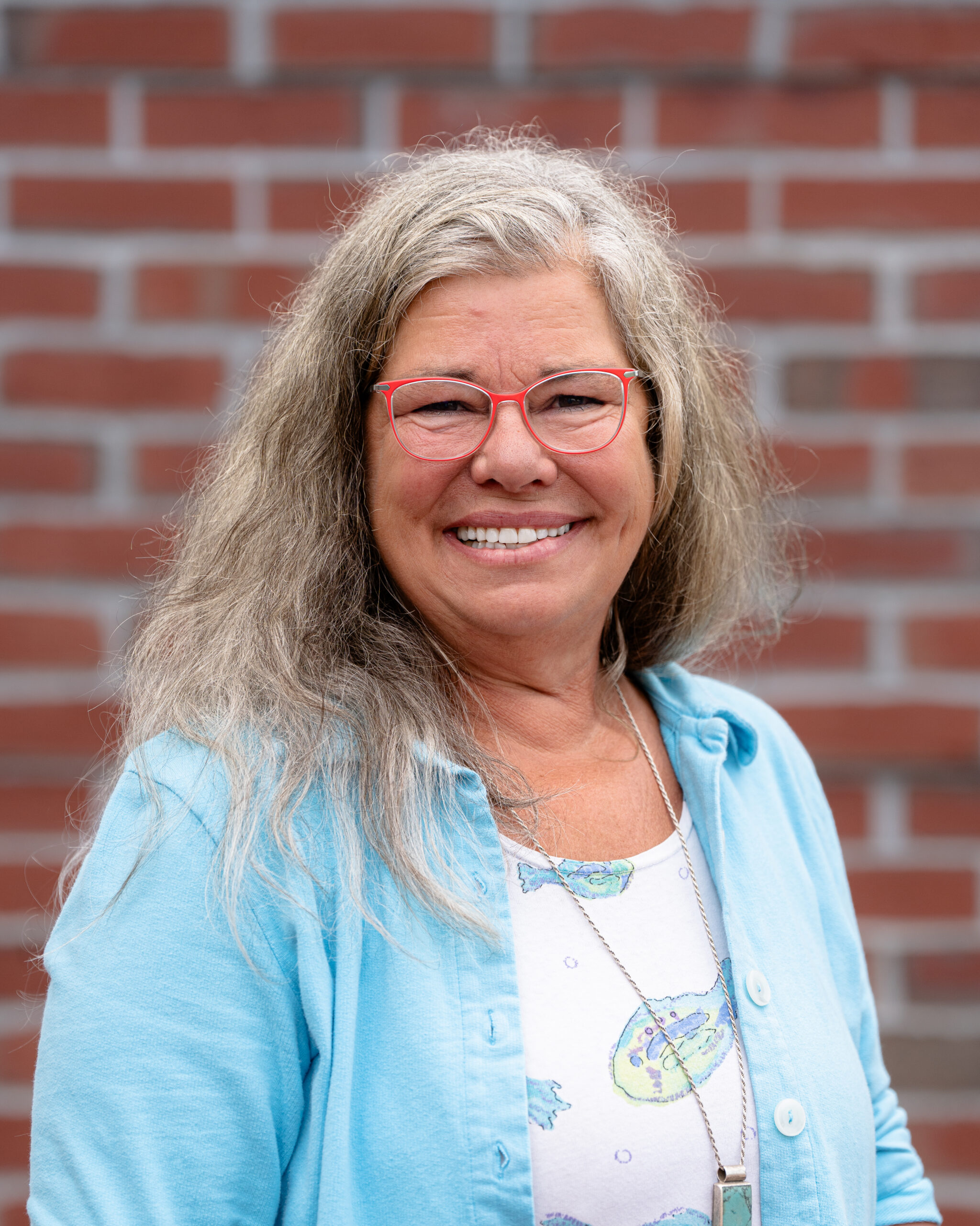 Kim, a white woman with curly gray hair. She wears red glasses and a blue jacket and stands in front of a red brick wall.