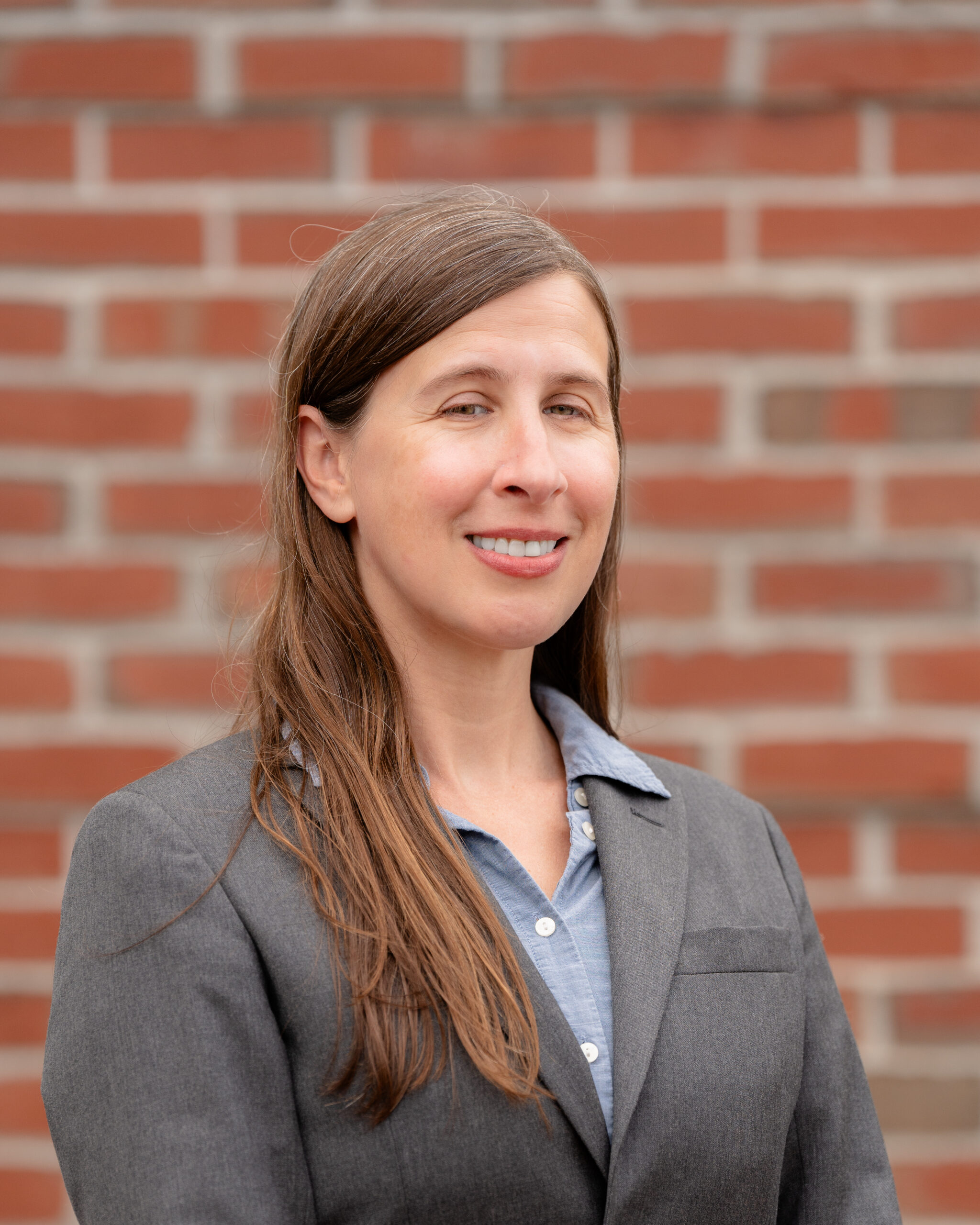 Lauren, a white woman with long brown hair. She wears a gray blazer and blue shirt.