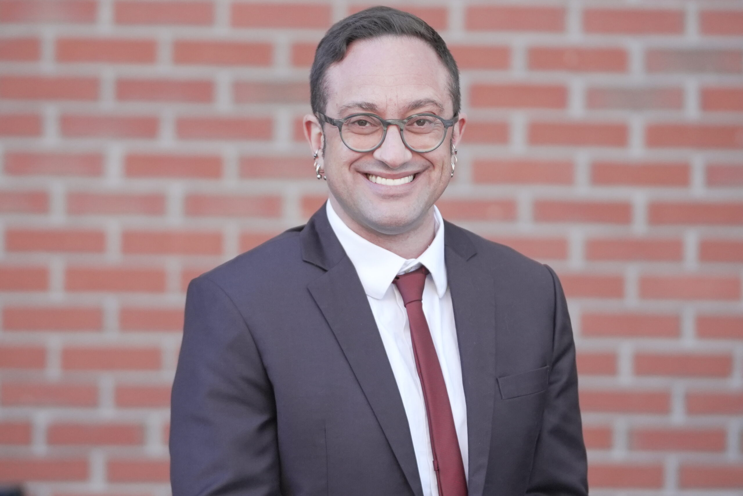 Lucas, a latino man with short brown hair and glasses. He wears a suitjacket and red tie.