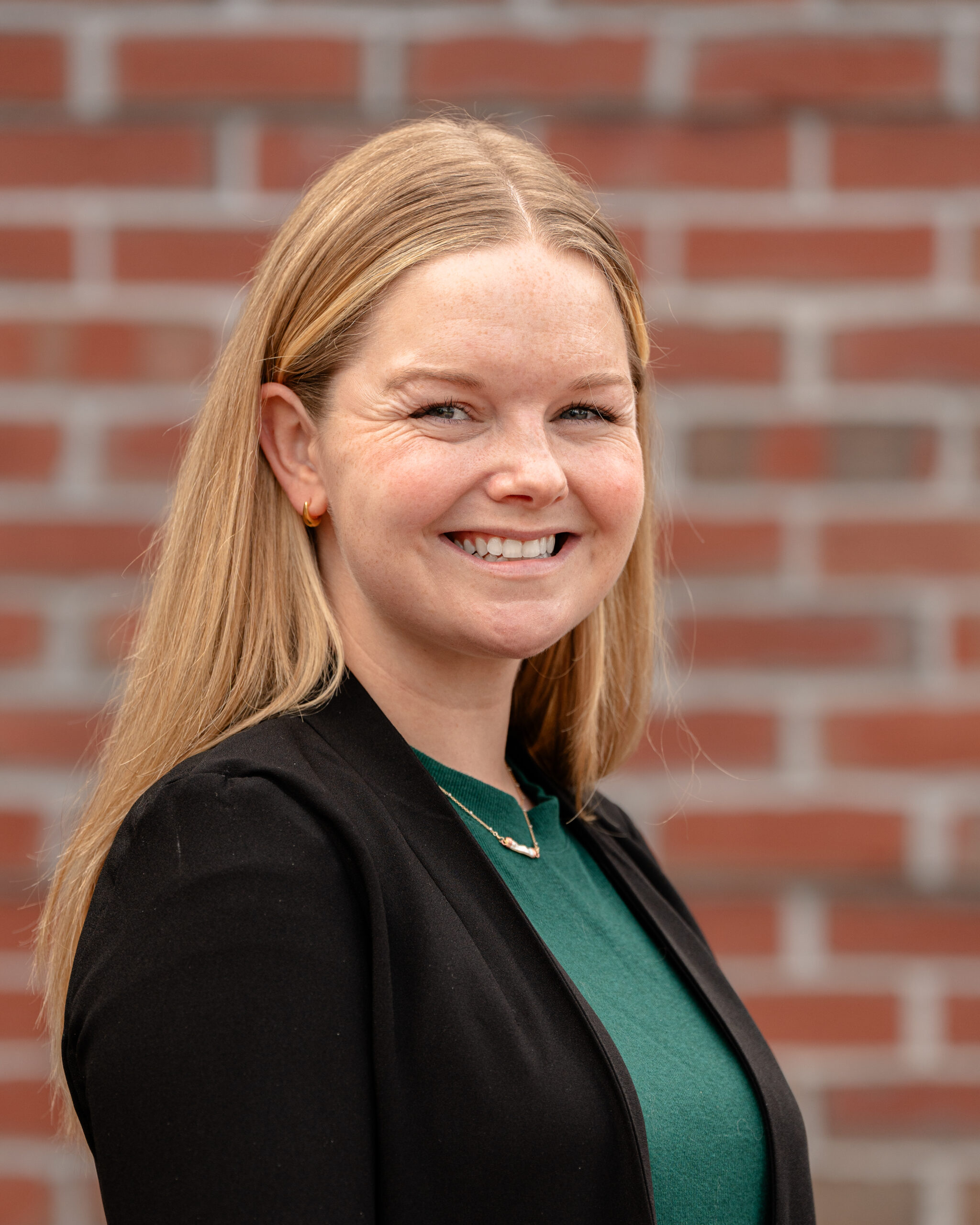 Megan, awoman with blonde hair smiles at the camera, wearing a green top and black blazer, standing in front of a blurred red brick wall.