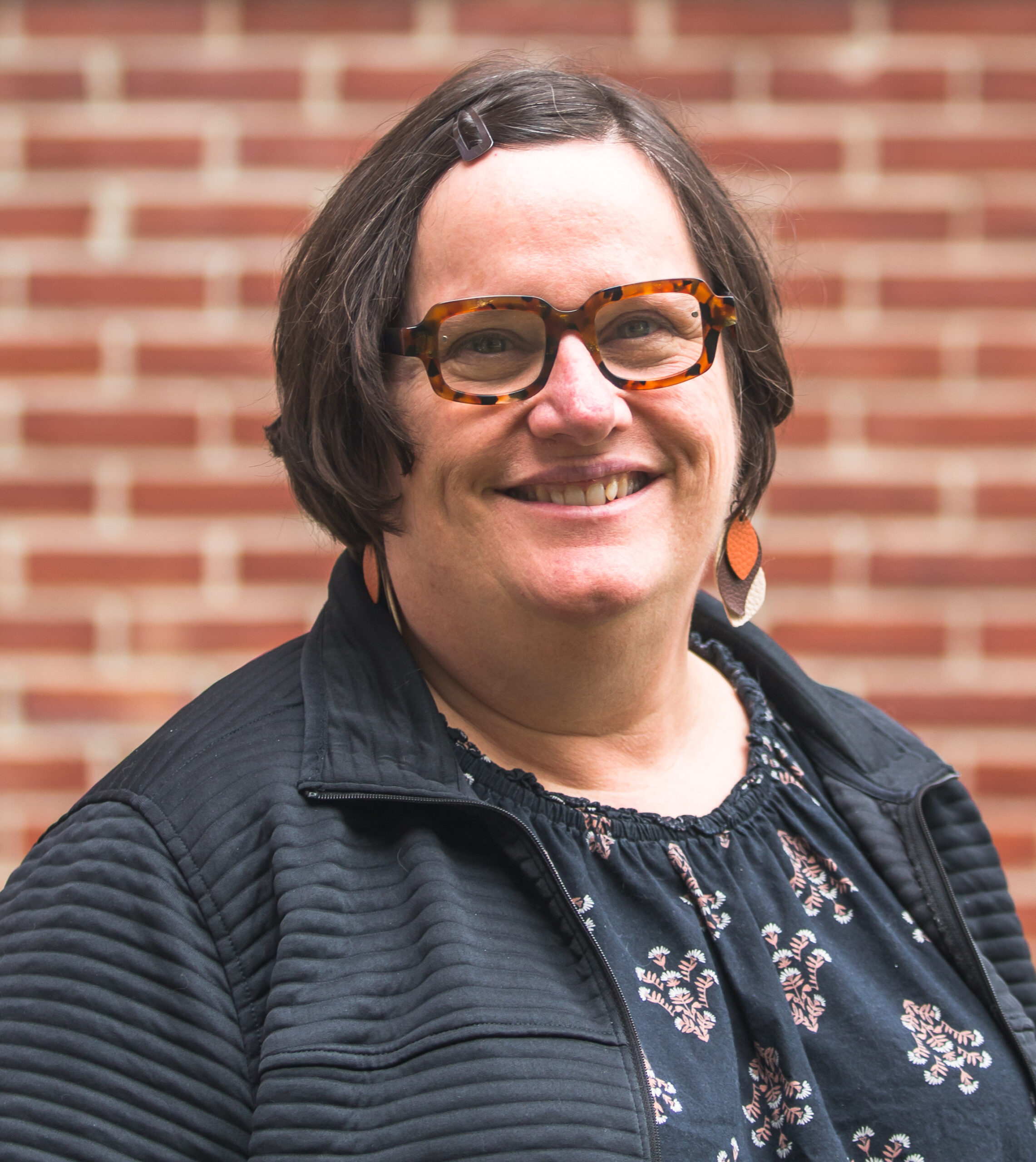 Michelle, a white woman with short brown hair and brown glasses. She wears long colorful earrings, a black jacket, and a floral shirt.