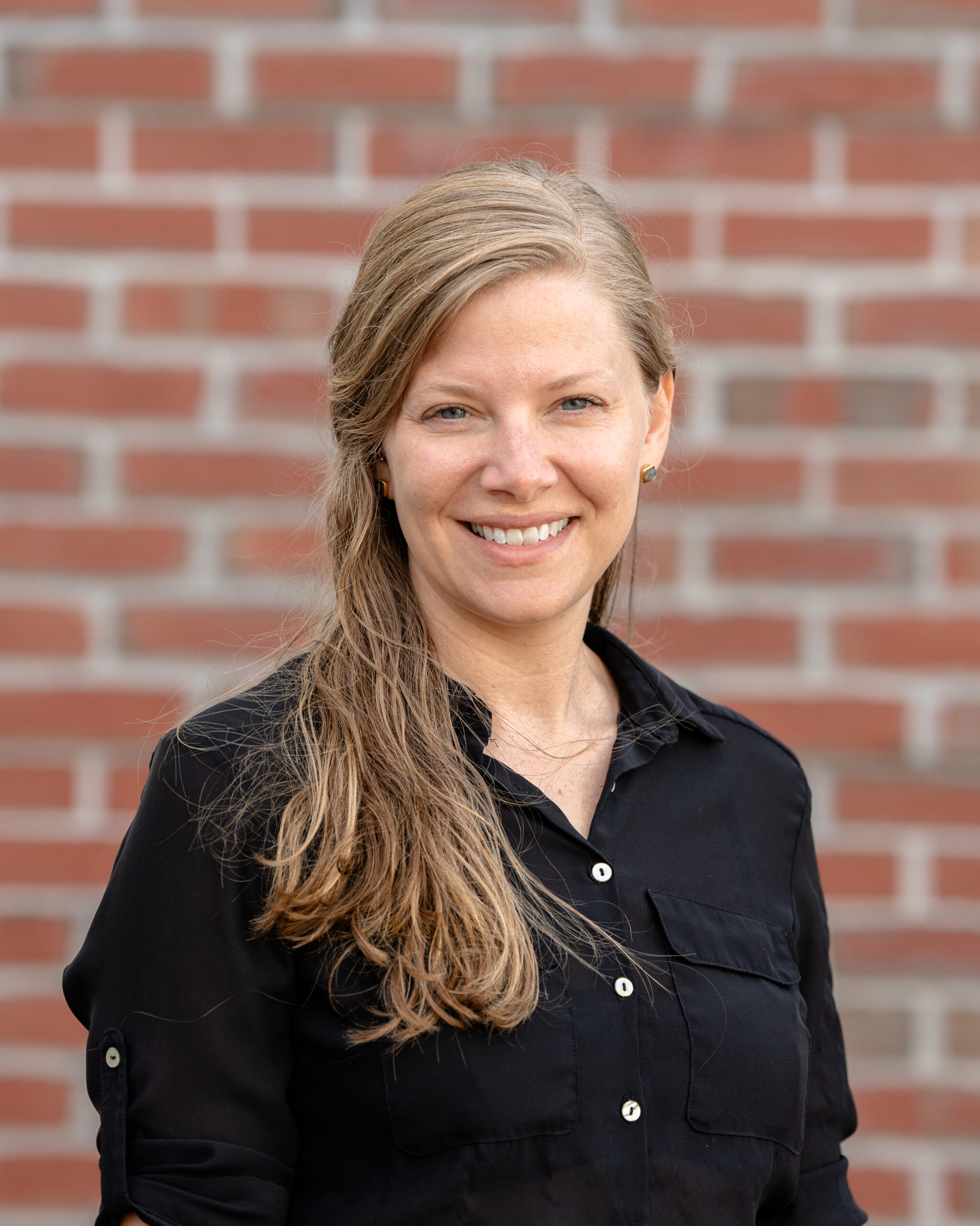 Riley, a white woman with dark blond hair, wearing a black shirt, is smiling.