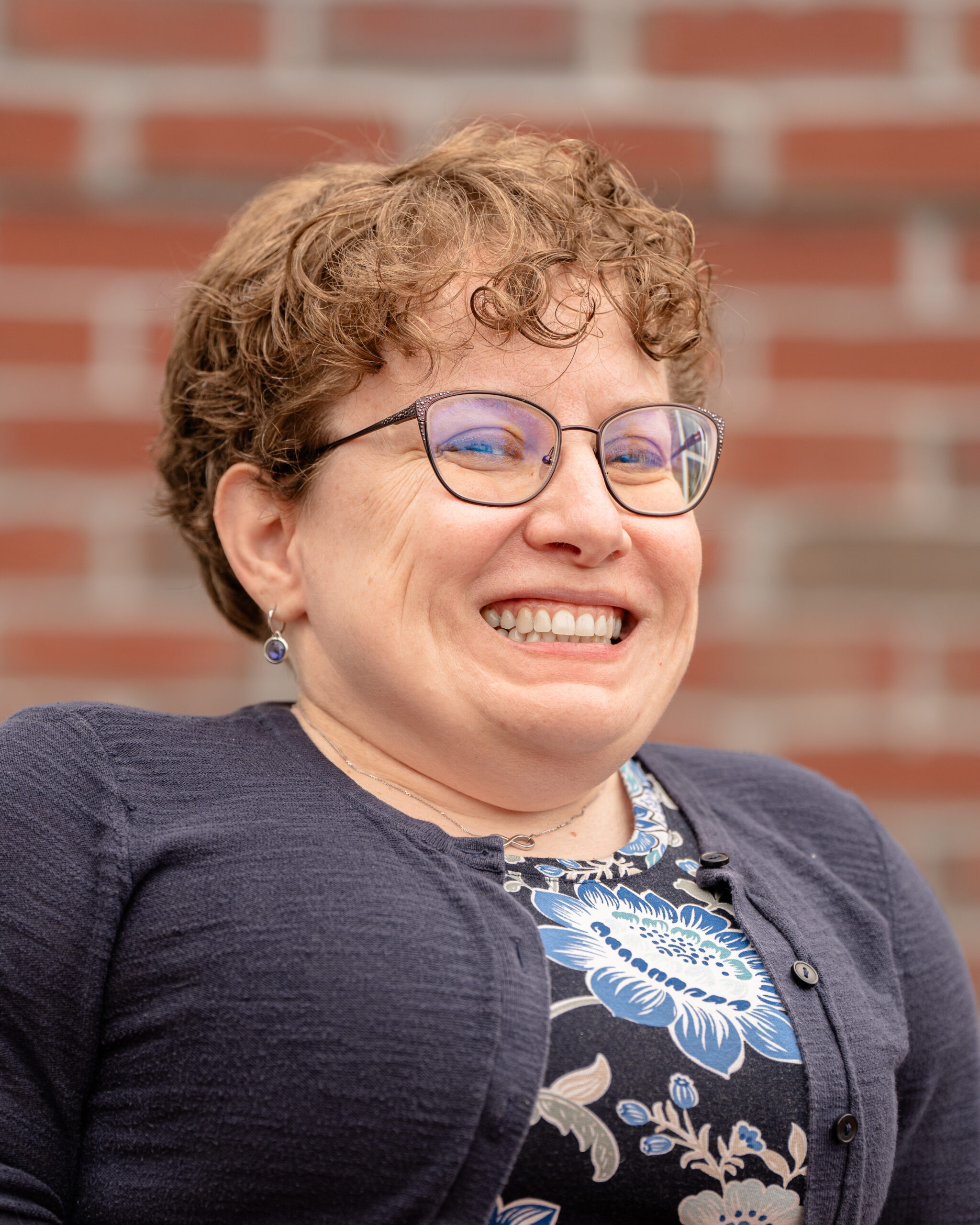 Sara, a woman with short, curly hair wearing glasses, earrings, a floral patterned dress, and a navy blue sweater.