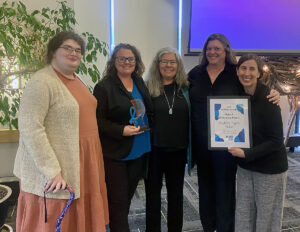 Group of five DRM staff standing in front of a colorful screen holding an award.