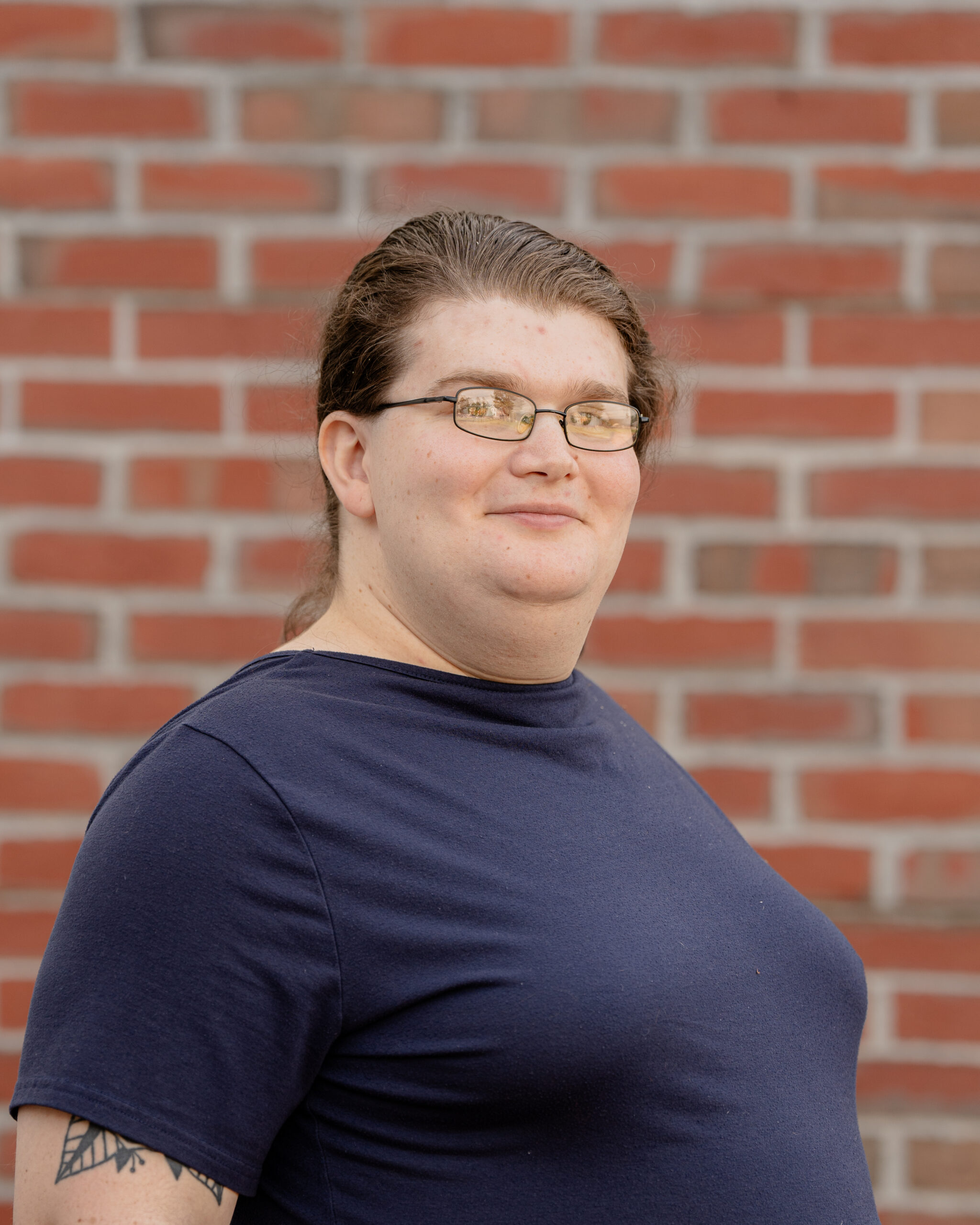 Headshot of Sammy-Ellie MacKinnon in front of a brick wall. She has tied back brown hair, square glasses, and is wearing a blue dress.