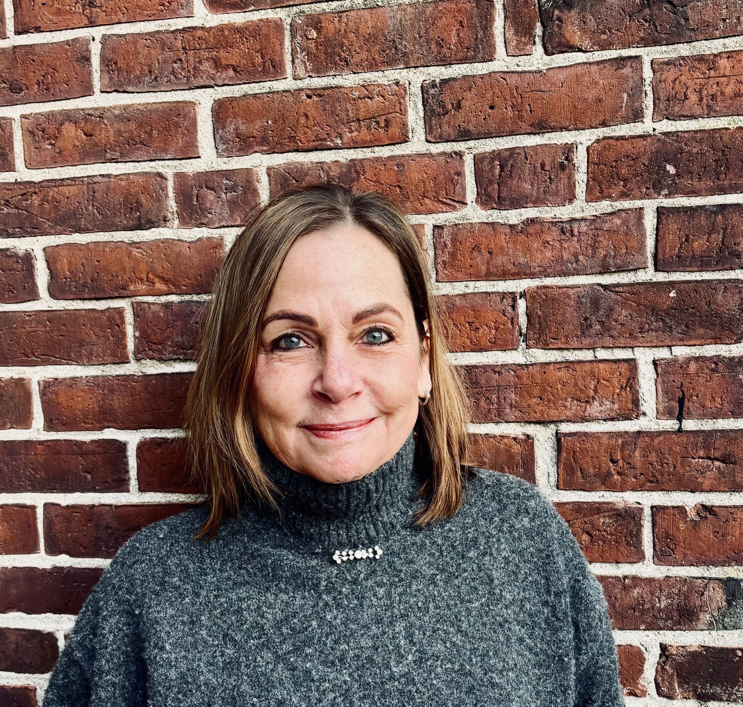 Susan, a white woman with short brown hair. She wears a gray sweater and stands in front of a brick wall.