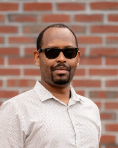 William, a black man with short brain hair and a short beard wears sunglasses and a light button down shirt.