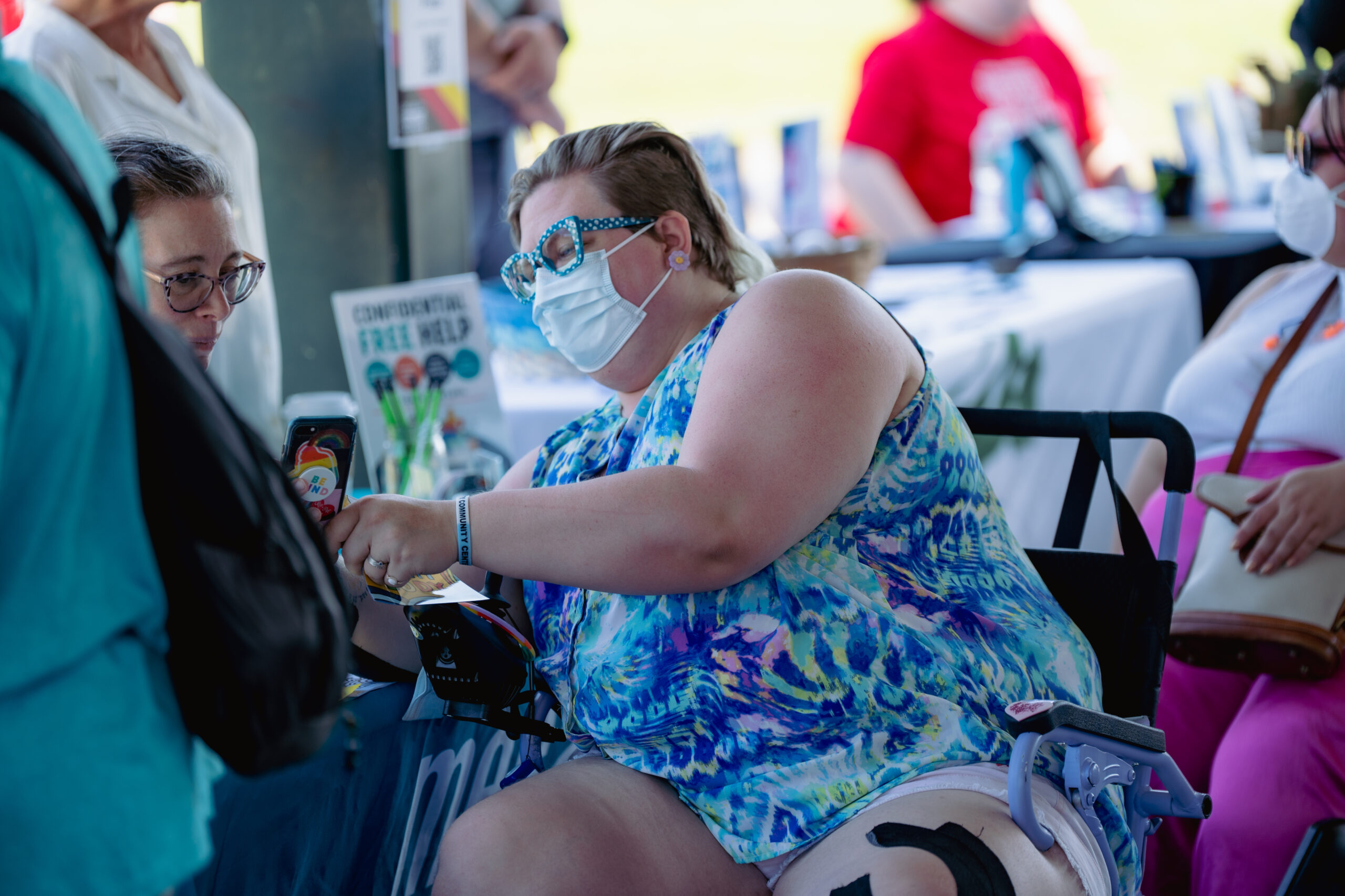 A person looks at items as they navigate in their power wheelchair. They wear a mask, blue glasses, and a colorful tank top.