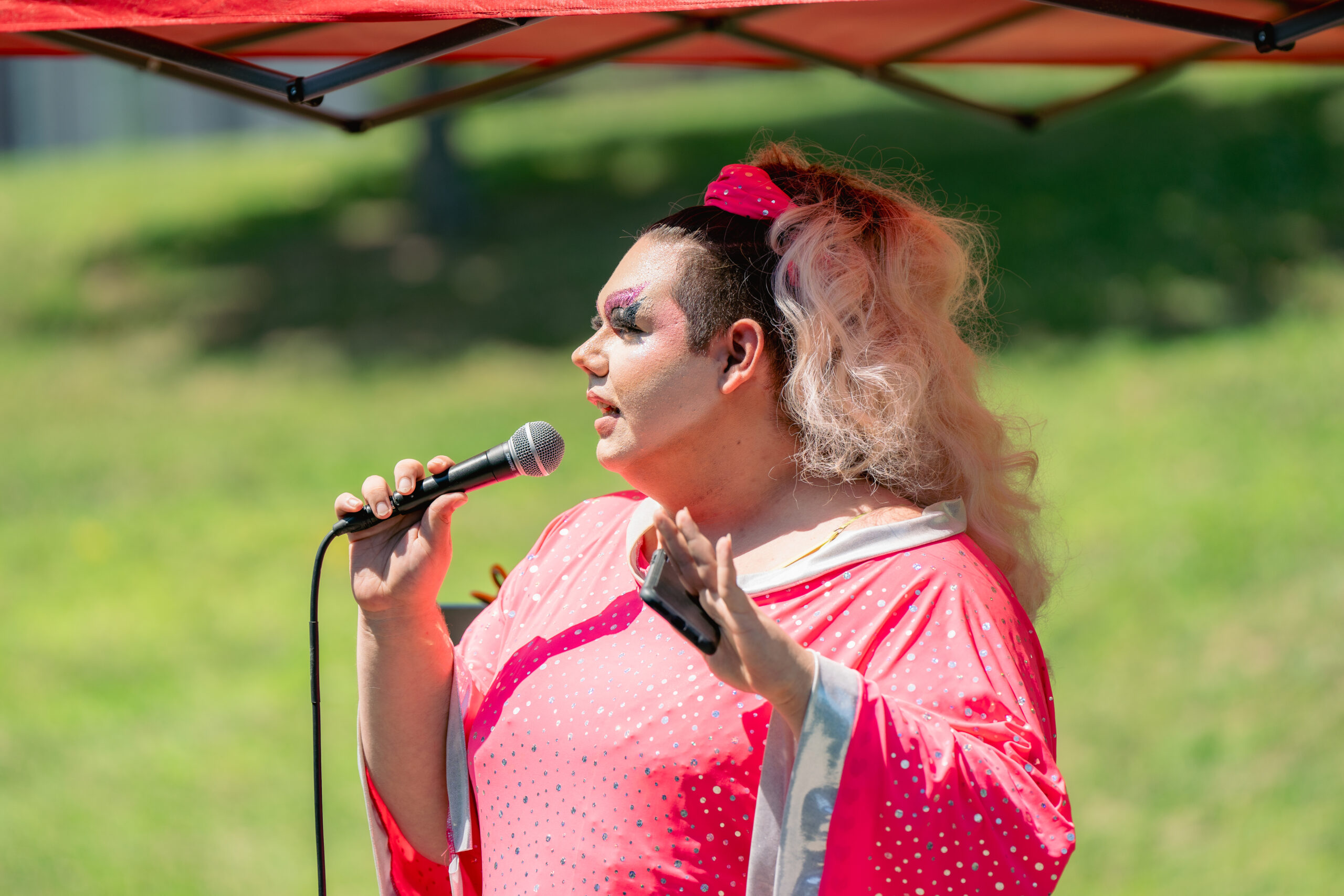 Person wearing pink dress with long blonde hair and elaborate face makeup holding a microphone.