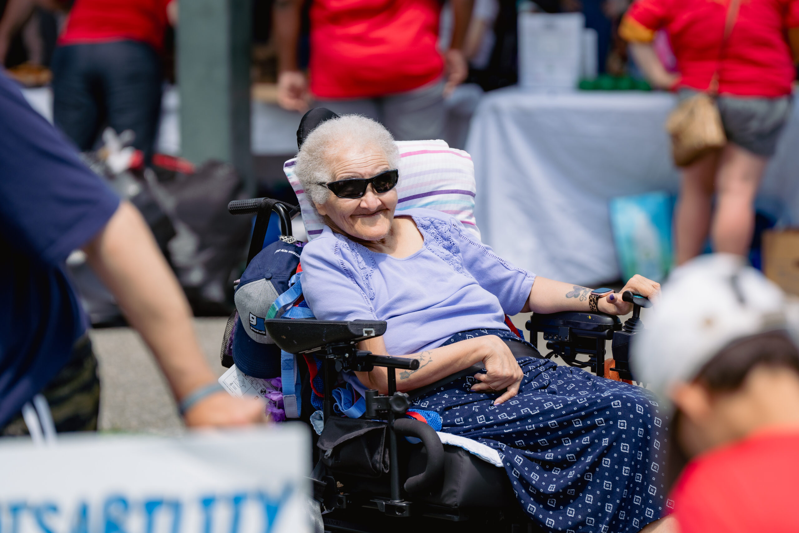 An older woman in power wheelchair. She wears sunglasses, a purple shirt, and a patterned skirt.