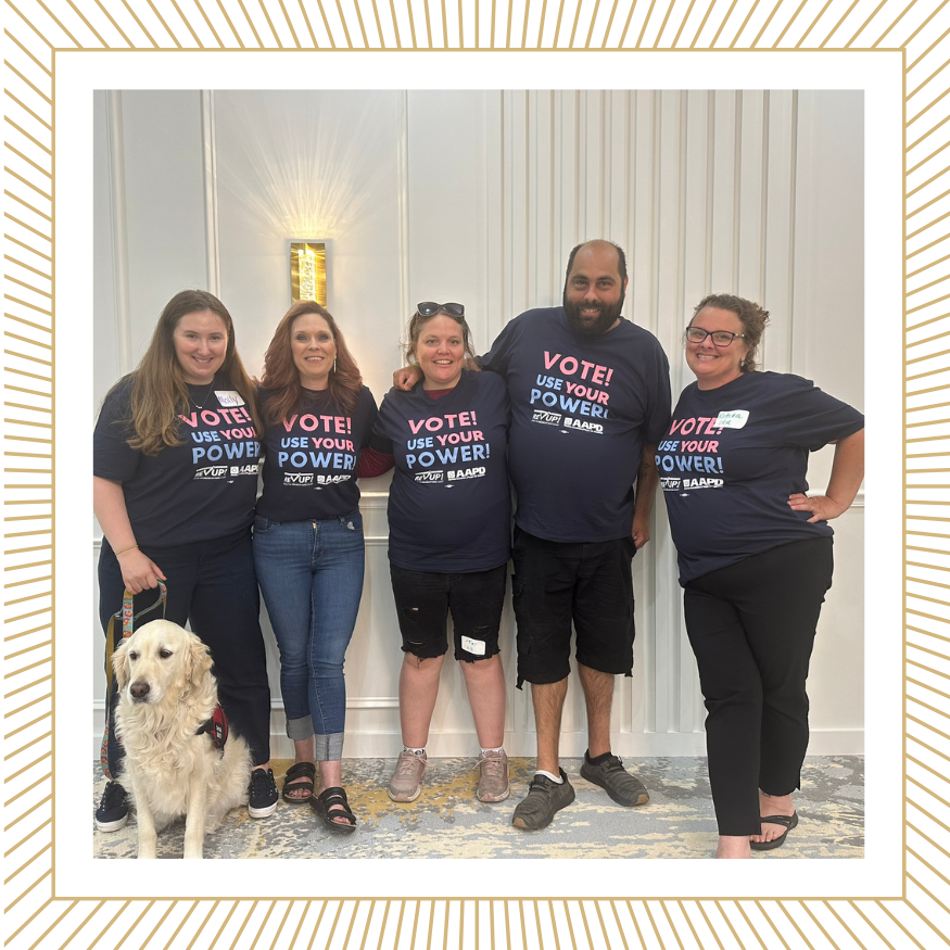 A group of 5 people and a golden retriever service dog stand in a group. They wear matching shirts that read Vote. Use Your Power. RevUp.