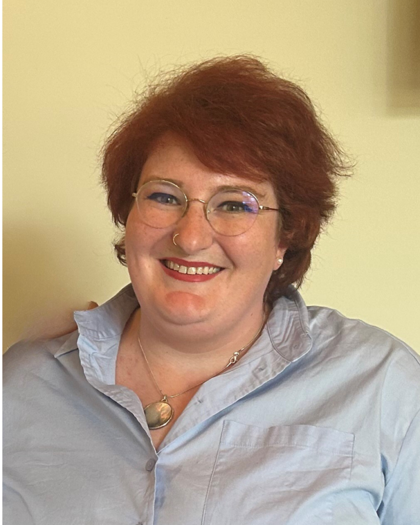 Kate, a white plus sized person, with red hair, glasses, and a blue button down shirt.