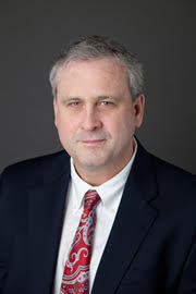 Christopher is a white man with short gray hair. He wears a suit, red tie, and white shirt.